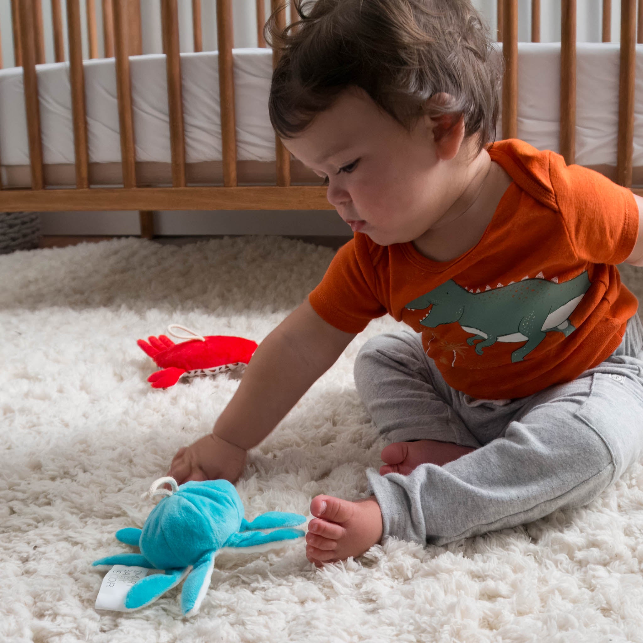 A baby sits on a fluffy white rug playing with plush characters of an octopus and crab.