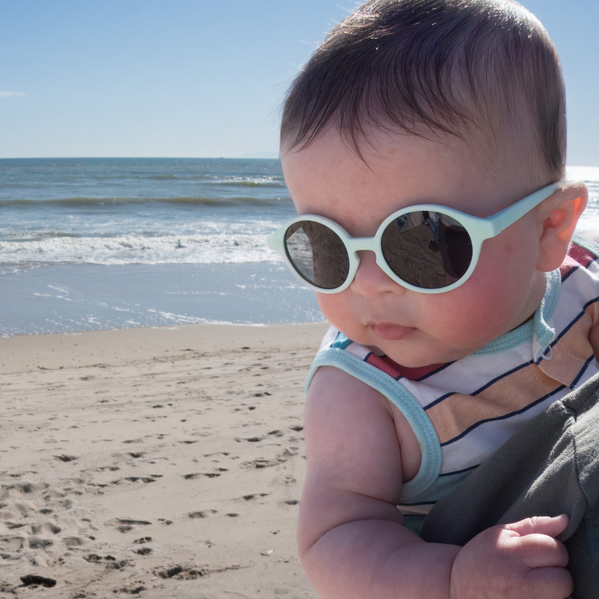 A baby is wearing light blue sunglasses while being held at the beach. 