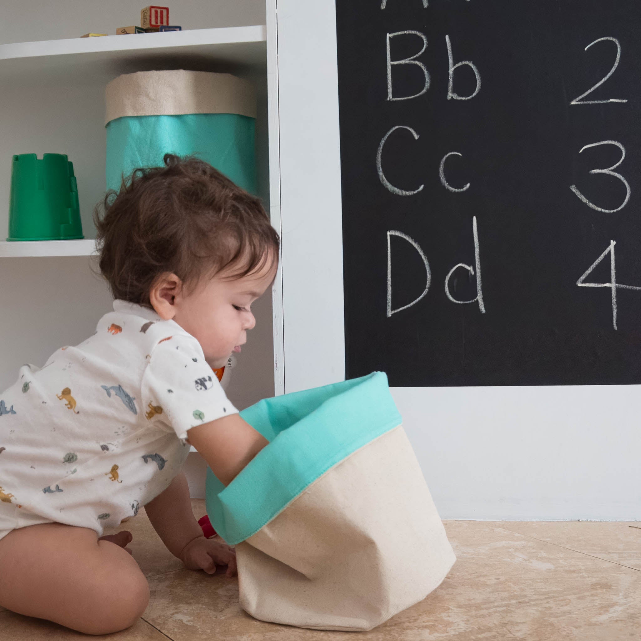 A baby reaching into a cloth storage basket that is beige on the outside and mint green on the inside. A chalkboard and shelf is in the background.