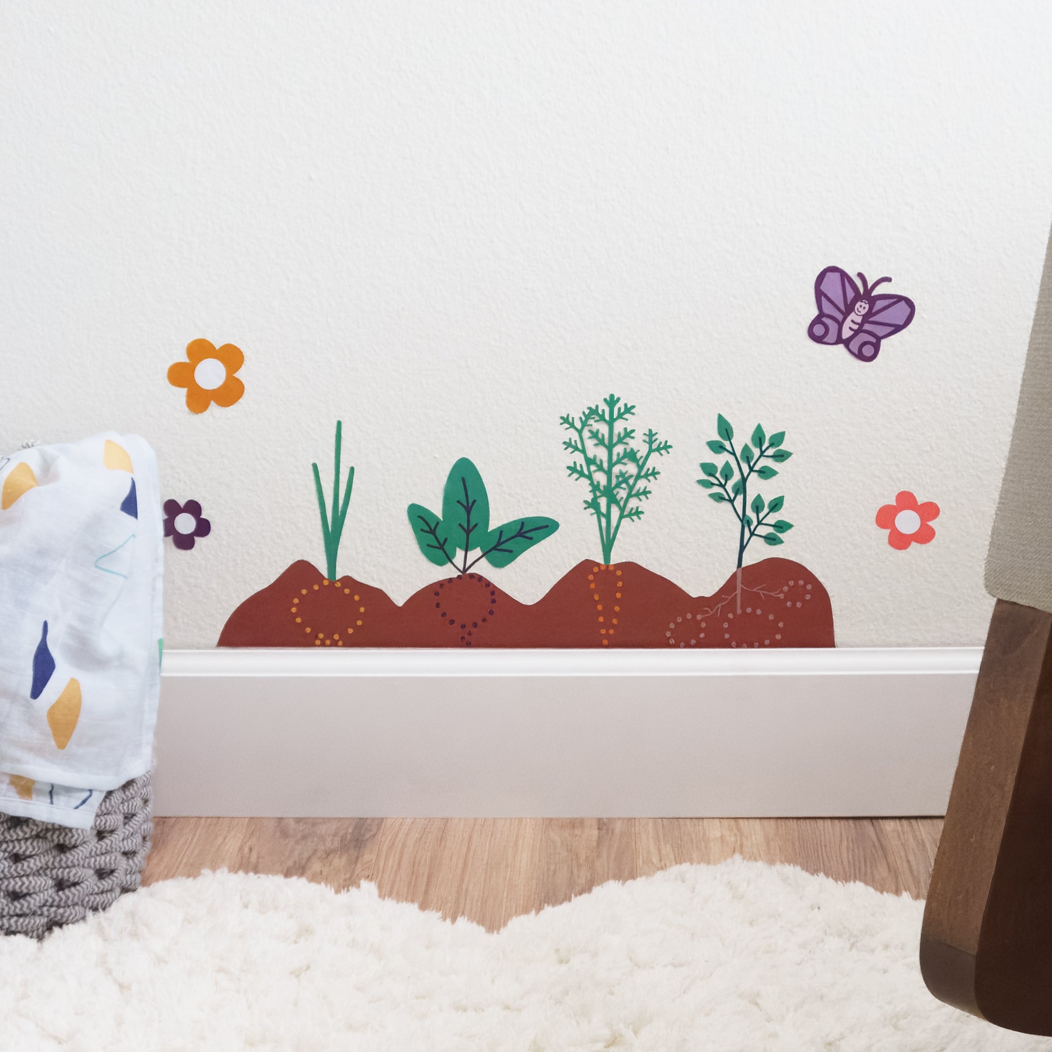 A close up of a wall art decal featuring a garden with an onion, beet, carrot and potato, surrounded by a few flowers and a butterfly. In front of the wall is a white, fluffy rug, a storage basket with blanket with triangle design and the edge of a rocking chair.