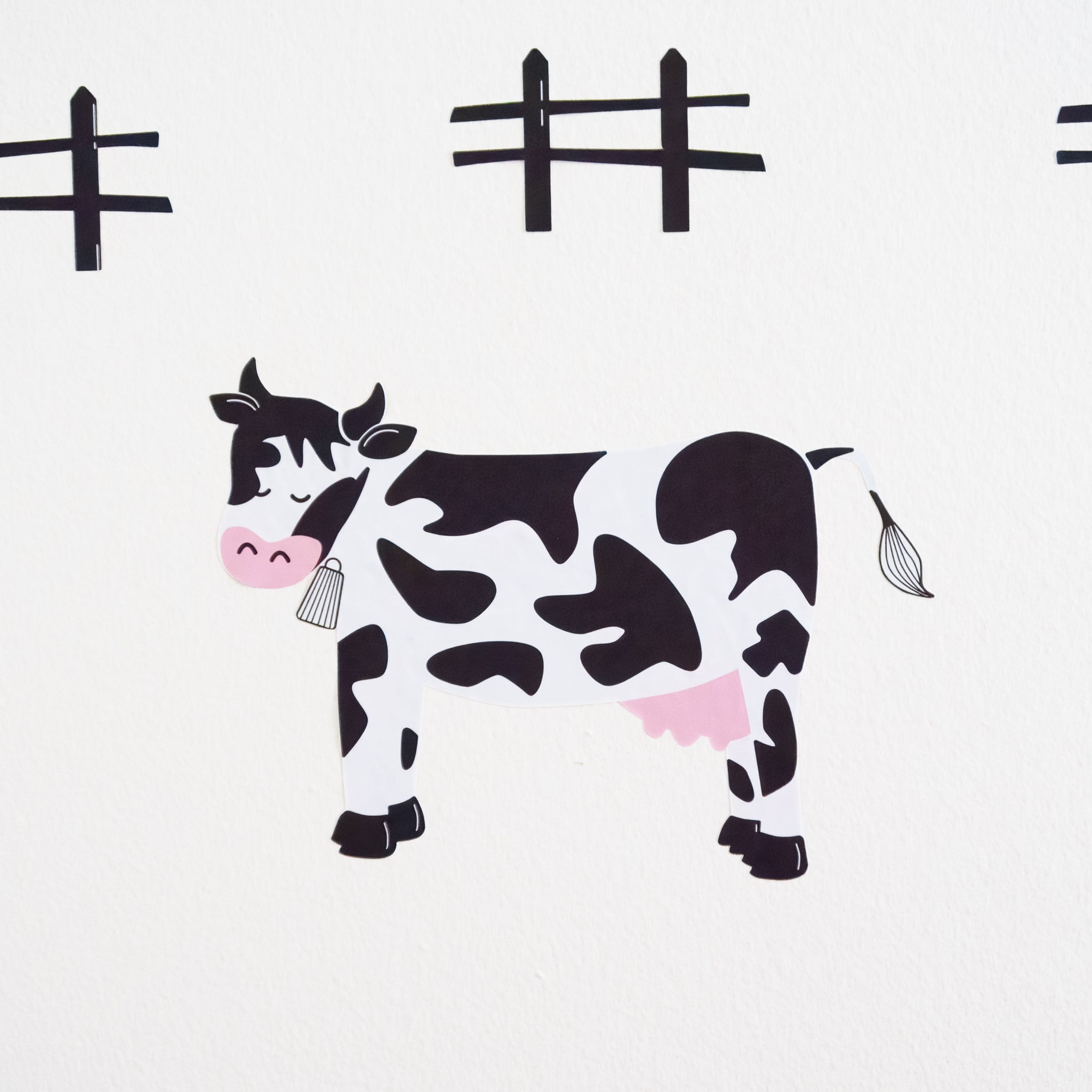 A black and white cow wall art with eyes closed, a cowbell and pink utter, with a few black fences in the background on a white wall.