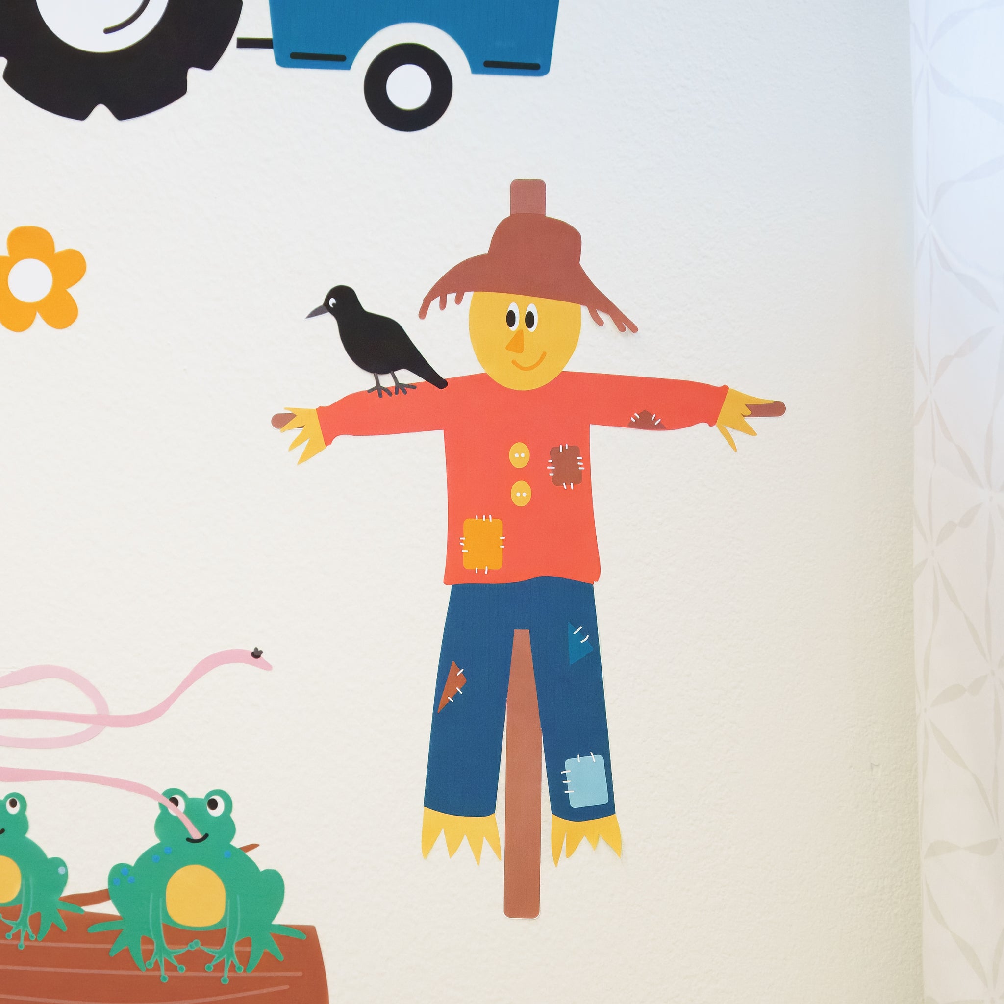 A close up of a scarecrow wall decal on a white wall with a red shirt, blue pants and a brown straw hat. He's looking to the left, and there is a black crow on his right arm. He's smiling.