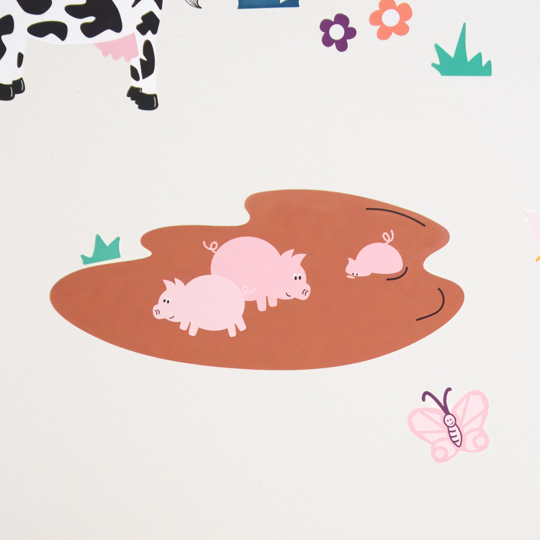 Wall decal of three pigs in a mud pit, one is half submerged. It is on a white wall with a butterfly, some flowers and grass around.