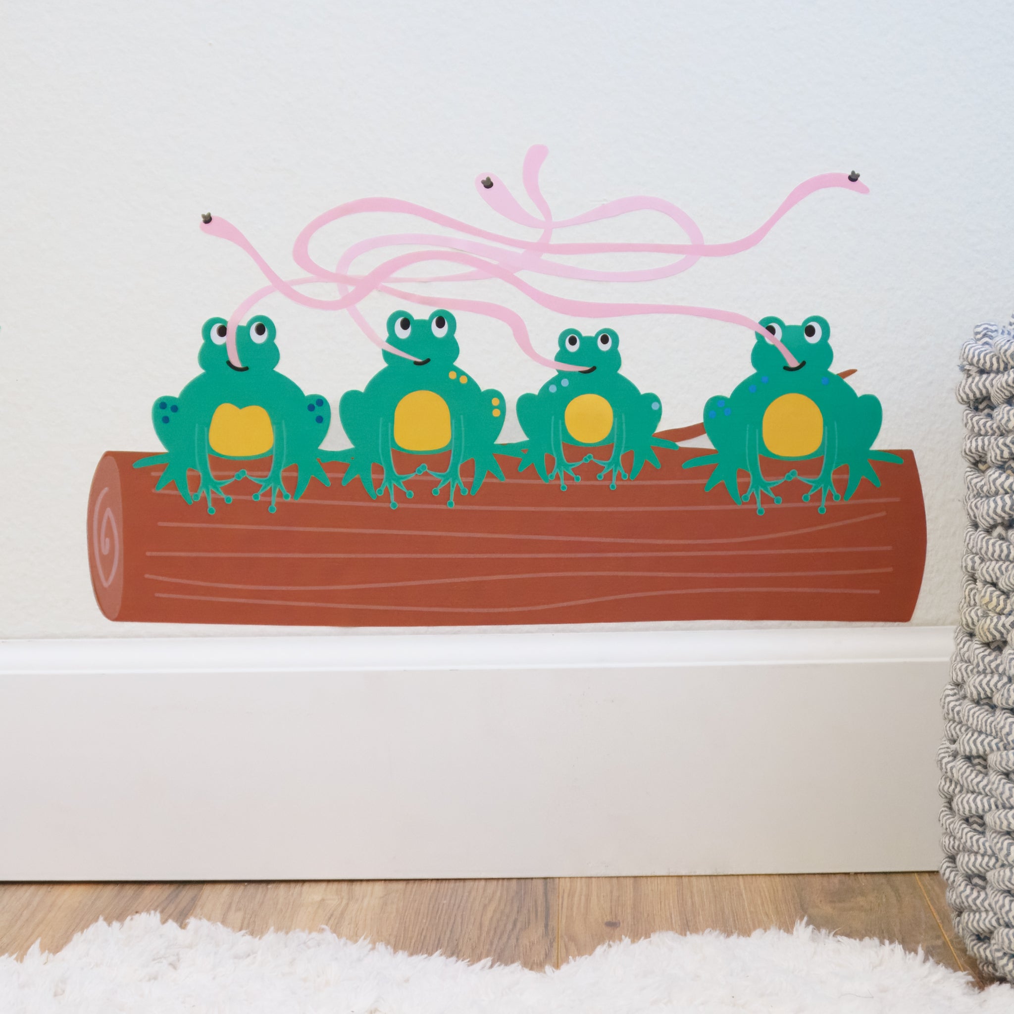 A close up of a wall decal of 4 green frogs with their tongues tangled trying to catch flies. They are sitting on a log. 