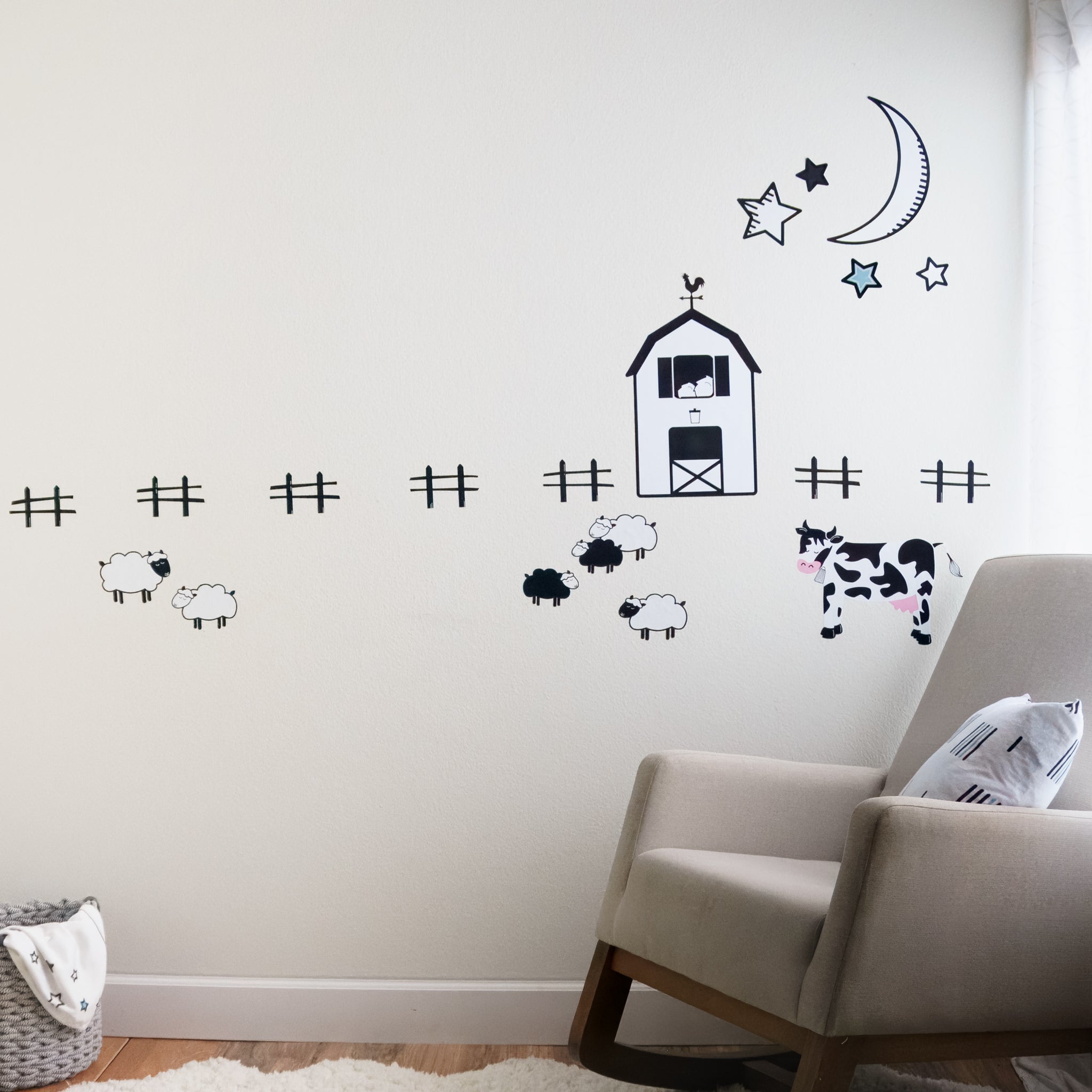 A farm scene on a white wall made with decals, including a black and white barn, sheep, fences, cow, moon and stars. In front is a rocking chair with throw pillow, white fluffy rug and storage basket. 