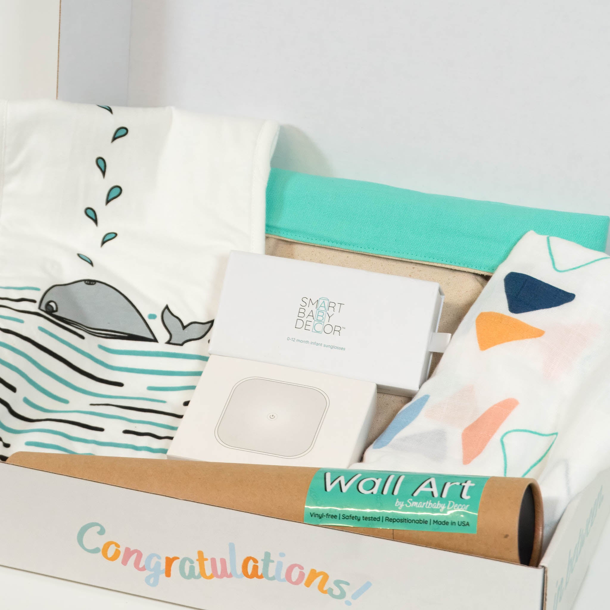 A box that says "congratulations" on the side is open and inside there is a baby burp cloth, night light, sunglasses box, wall art, blanket and storage basket.