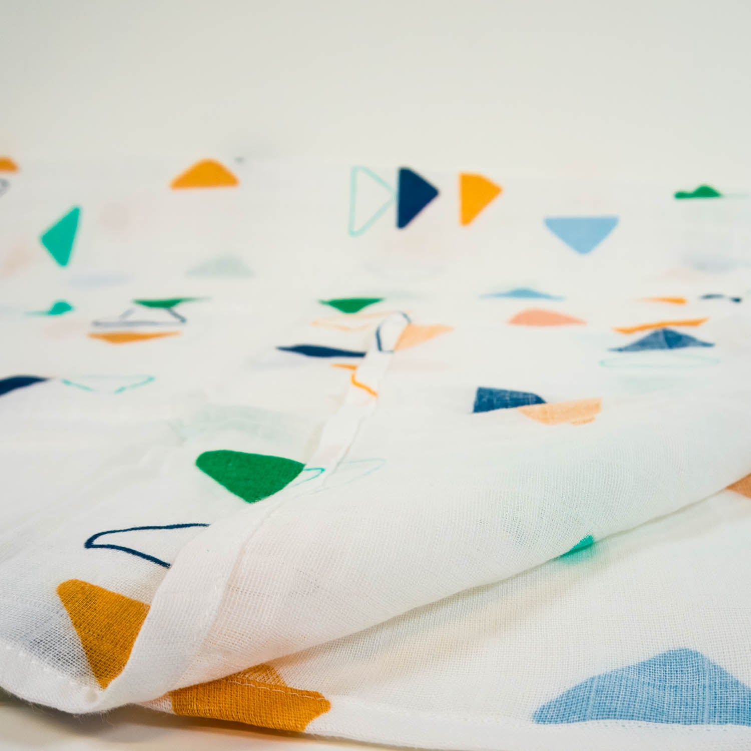 A close up of a white muslin blanket in white with a multicolored triangle pattern, with a corner flipped over.