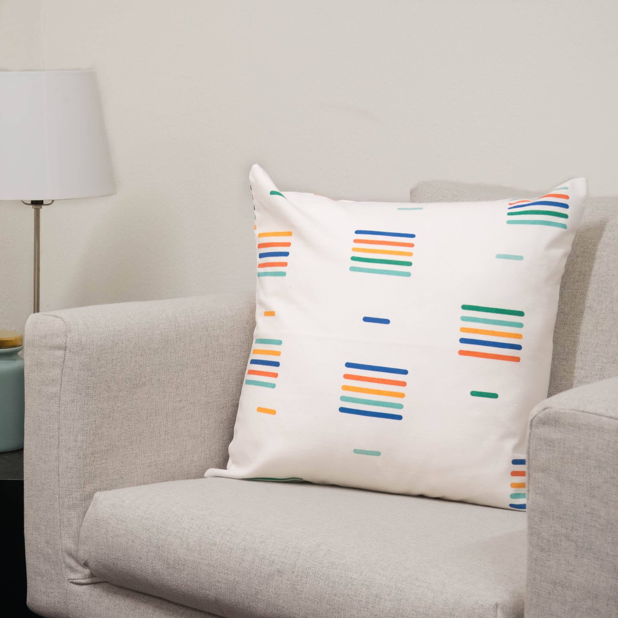 A chair with a throw pillow on top. The throw pillow is white with a linear repeated pattern in different bright colors. 