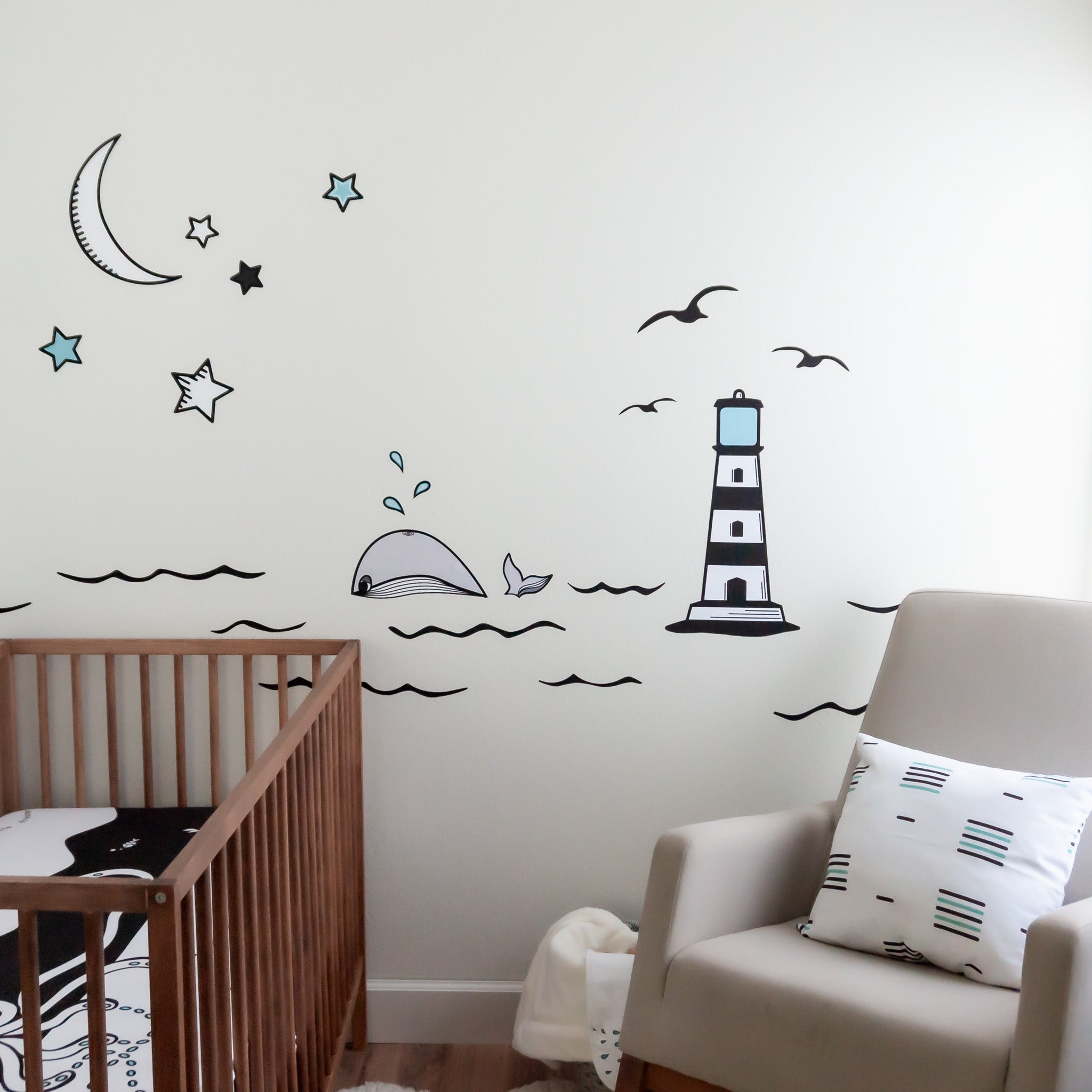 A nursery with a rocking chair on the right, wooden crib on the left with a black and white crib sheet. The wall has an ocean mural with a lighthouse, whale, moon and stars. 