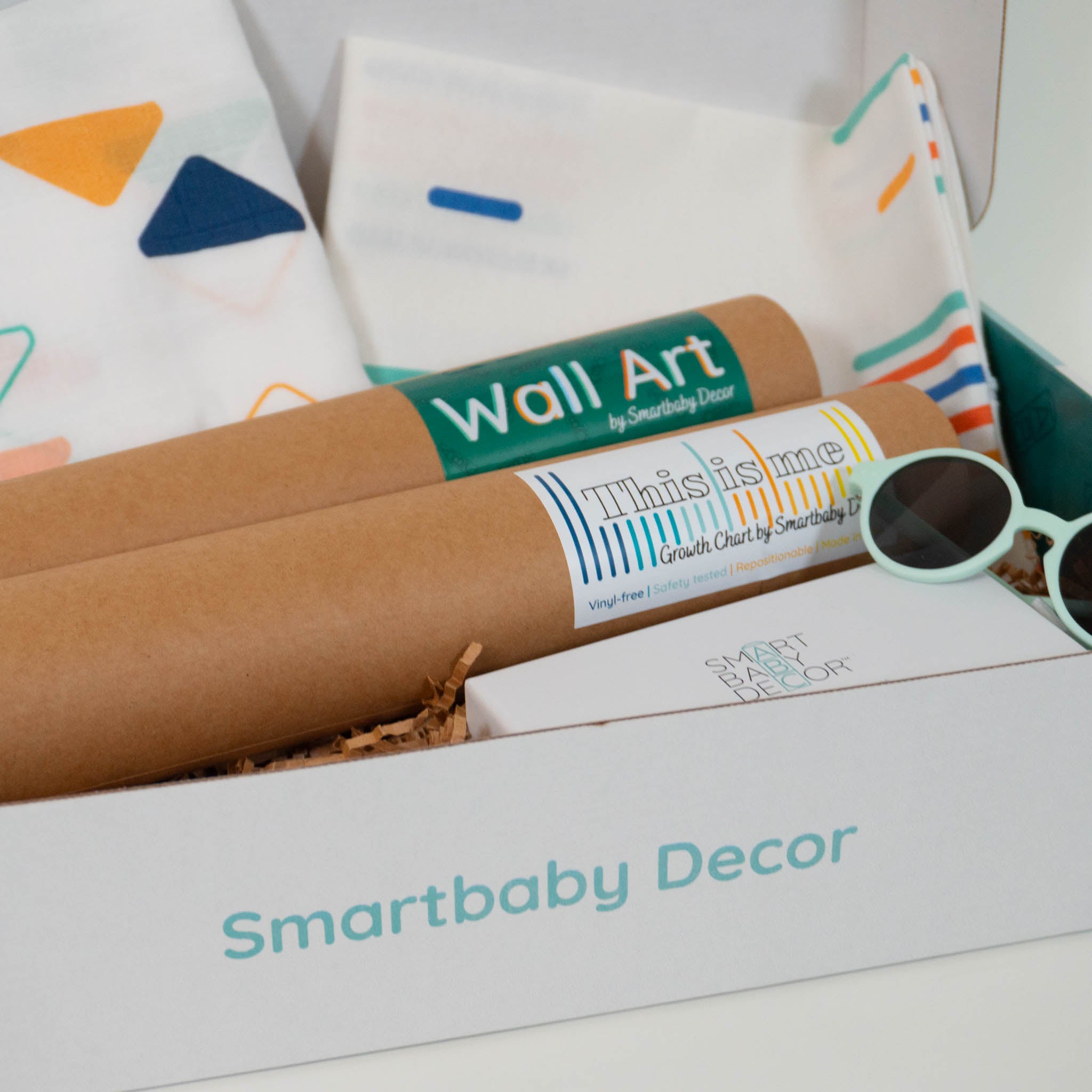 A closeup of the inside contents of Smartbaby Decor's Ready for Color Deluxe Box showing a pair of green infant sunglasses, 2 paper tubes of wall art, a blanket with a colorful triangle pattern and a throw pillow cover with a multicolored stripe pattern.