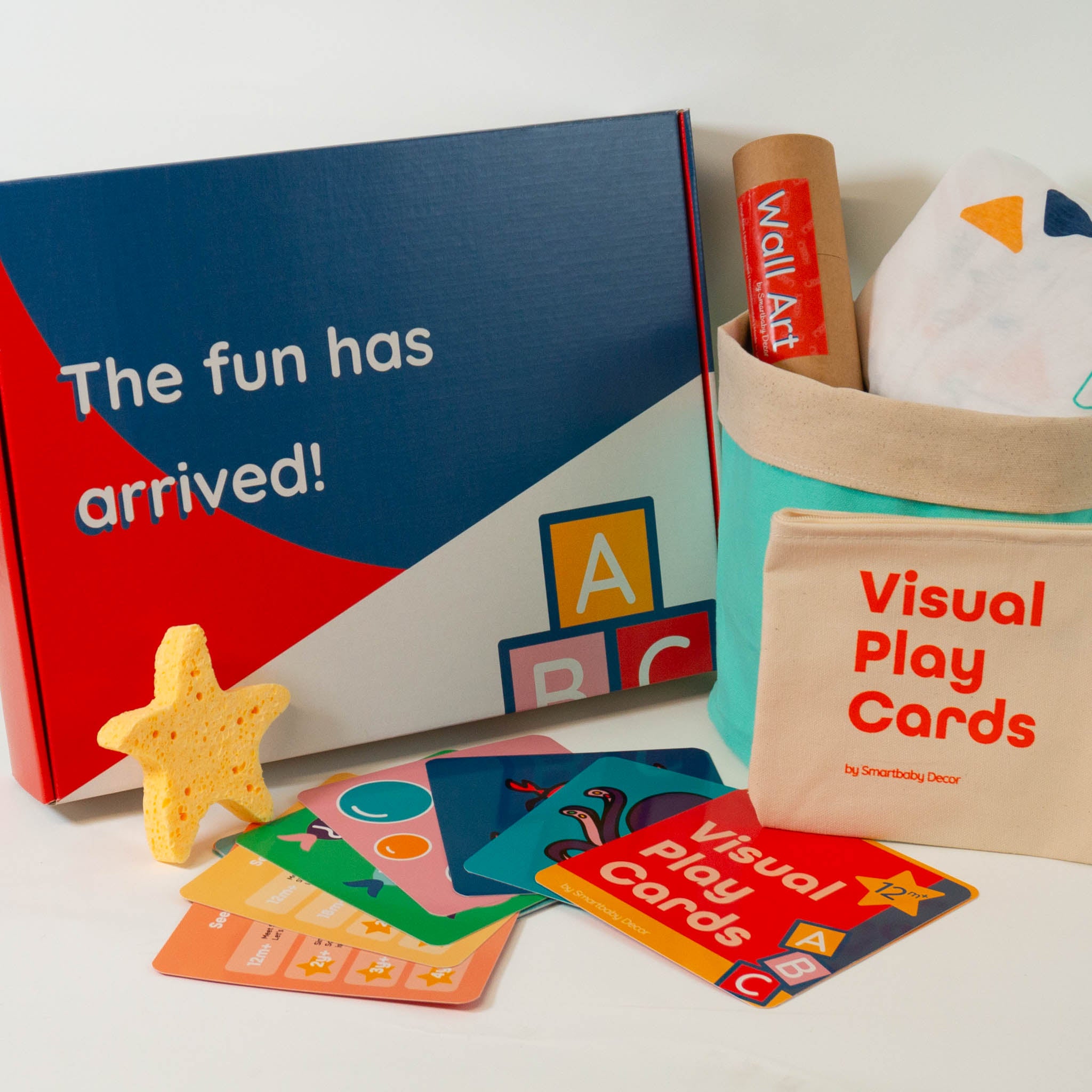 The outside of the Smartbaby Decor Deluxe Edition See Play Learn box is shown on the left reading "The fun has arrived!". On the right is a canvas storage basket in mint and beige, with a paper tube of wall art and a white crib sheet with triangle pattern inside. In front there is a set of colorful toddler activity cards with carrying case and a yellow starfish bath sponge.