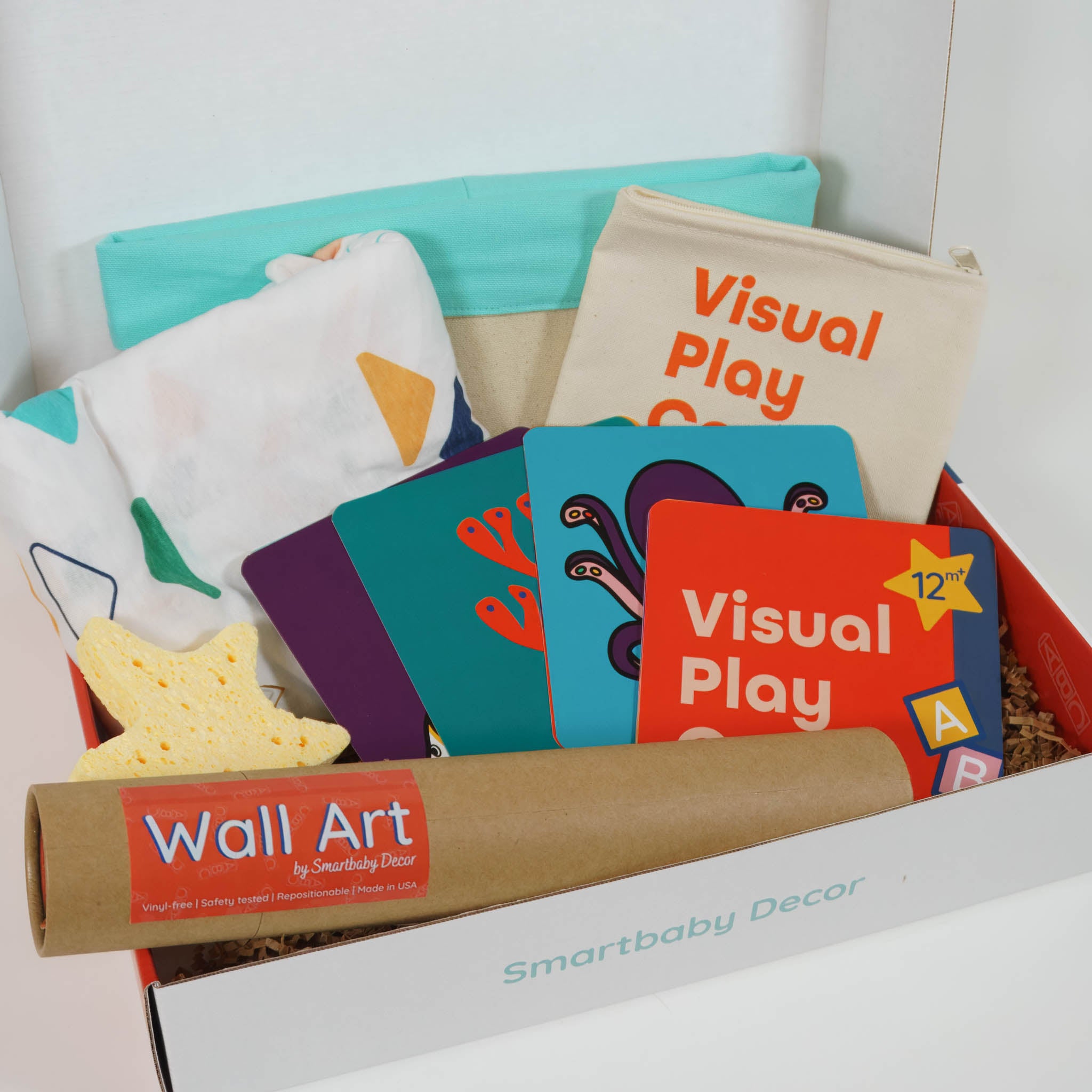 Inside contents of the Smartbaby Decor See Play Learn Deluxe Edition box showing a white crib sheet with multicolored triangle pattern, activity cards with carrying case, star bath sponge in yellow, a paper tube containing wall decals and a mint and beige storage basket.