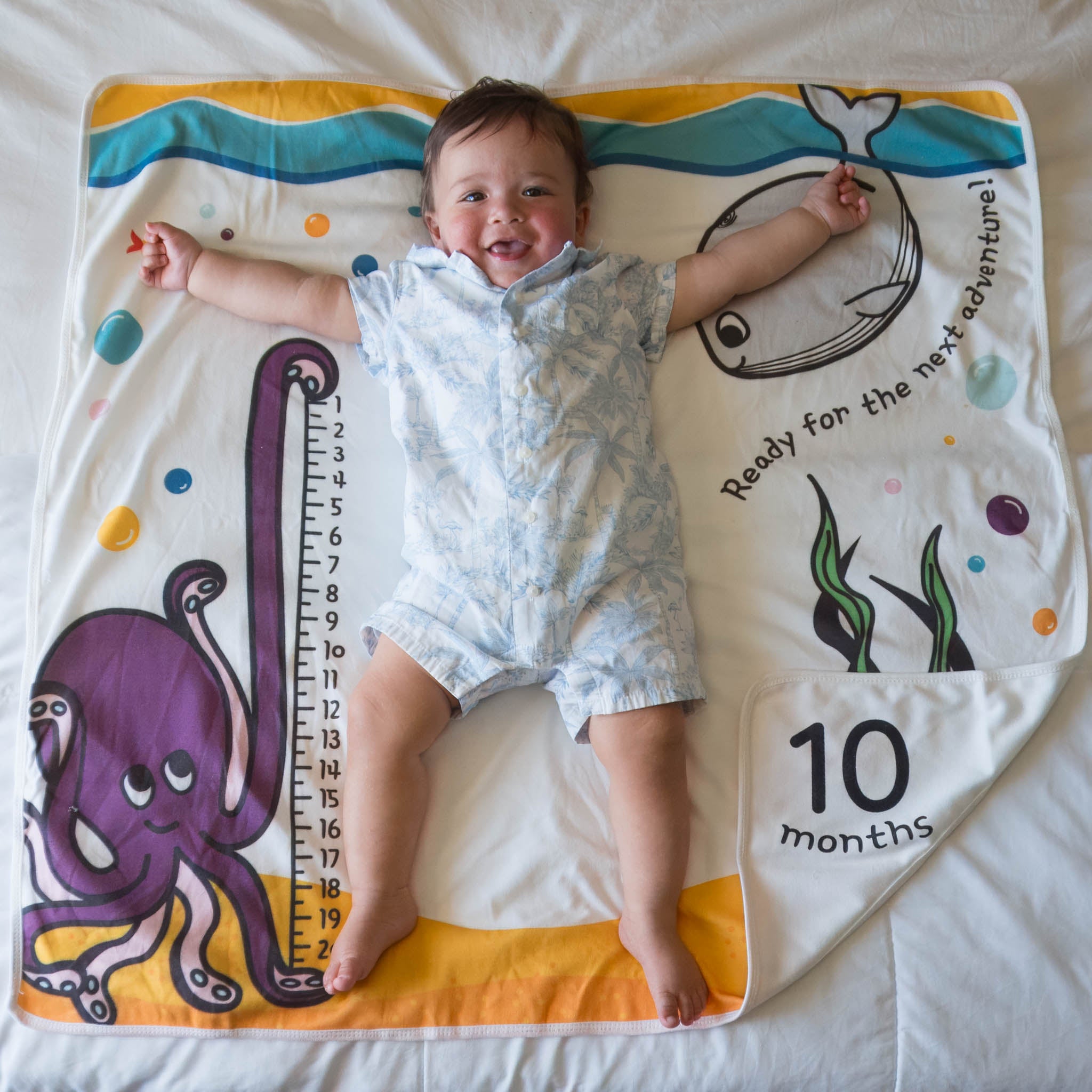 A baby lies with arms and legs out on a towel which has an ocean scene printed. The corner reads "10 months".