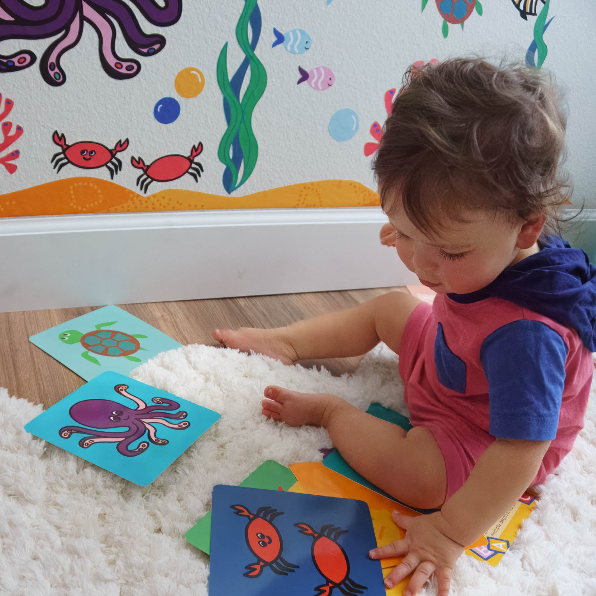 A toddler sits on a rug with colorful activity cards in an ocean theme, that match the wall mural on the wall in front of him.