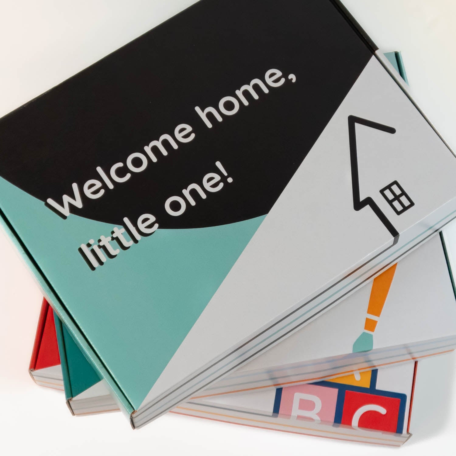 An aerial view of a stack of 3 gift boxes. The top one is black and mint with the phrase "welcome home, little one!". The other boxes are partially visible.