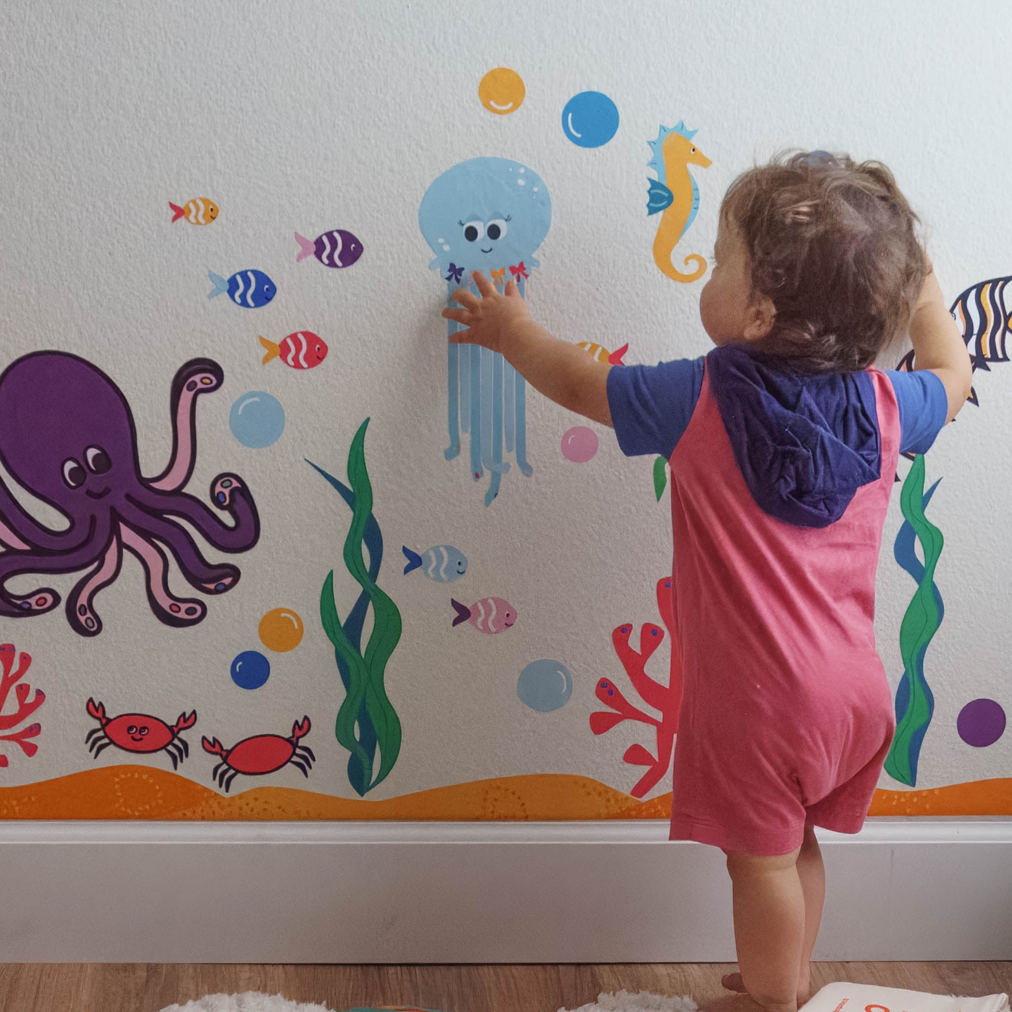 A toddler standing and touching a colorful ocean wall mural.