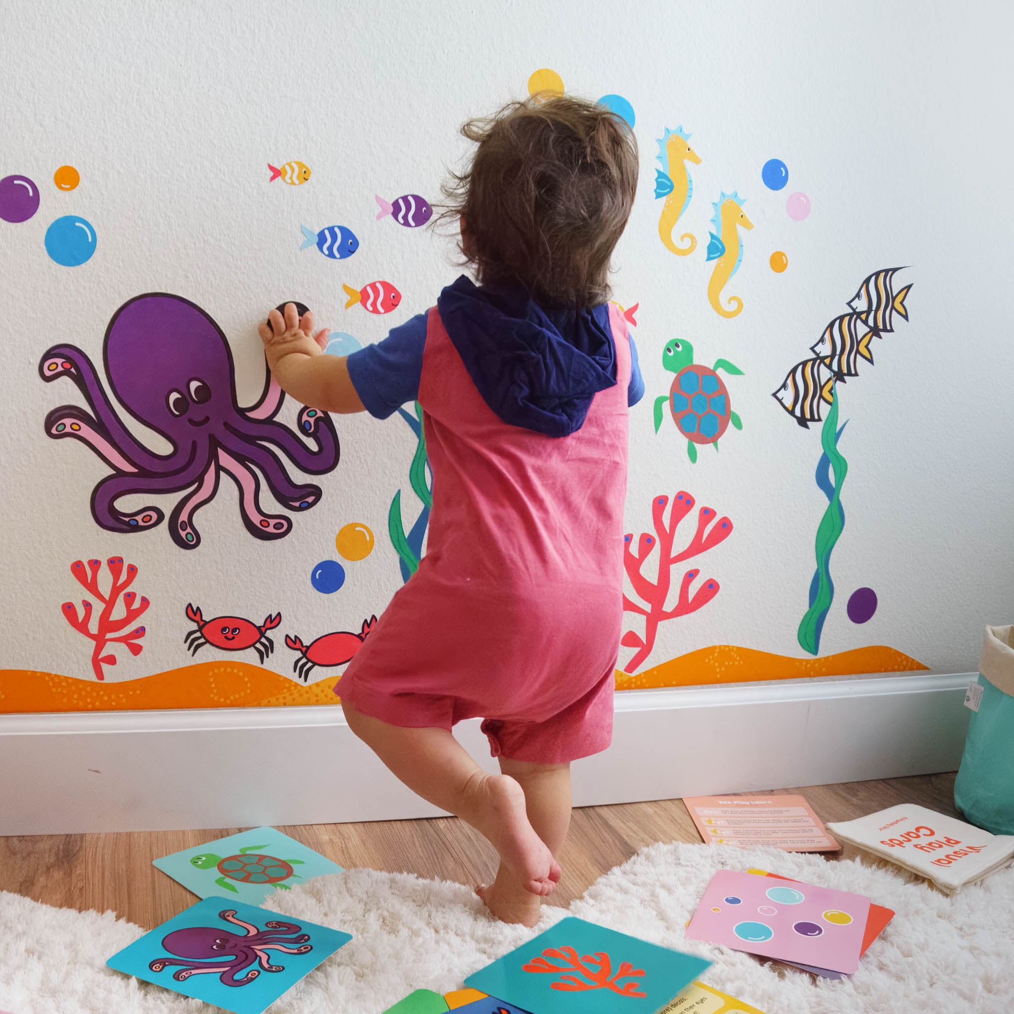 A toddler standing and touching an ocean wall mural with matching activity cards spread across the floor around him.