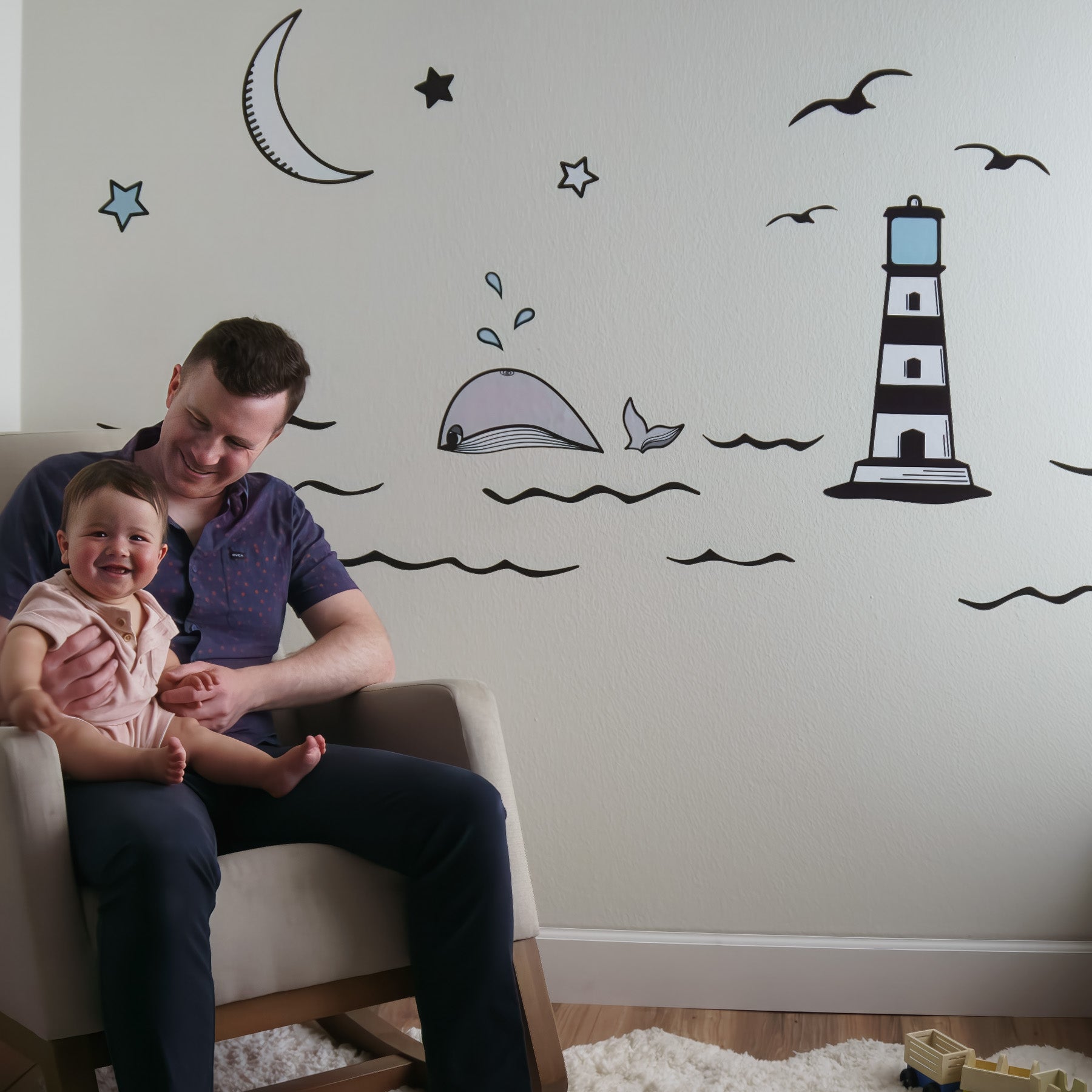 A wall mural of an ocean scene in the background with a dad and baby sitting in a rocking chair in front. The mural is black and white, the baby is laughing, and the dad is smiling.