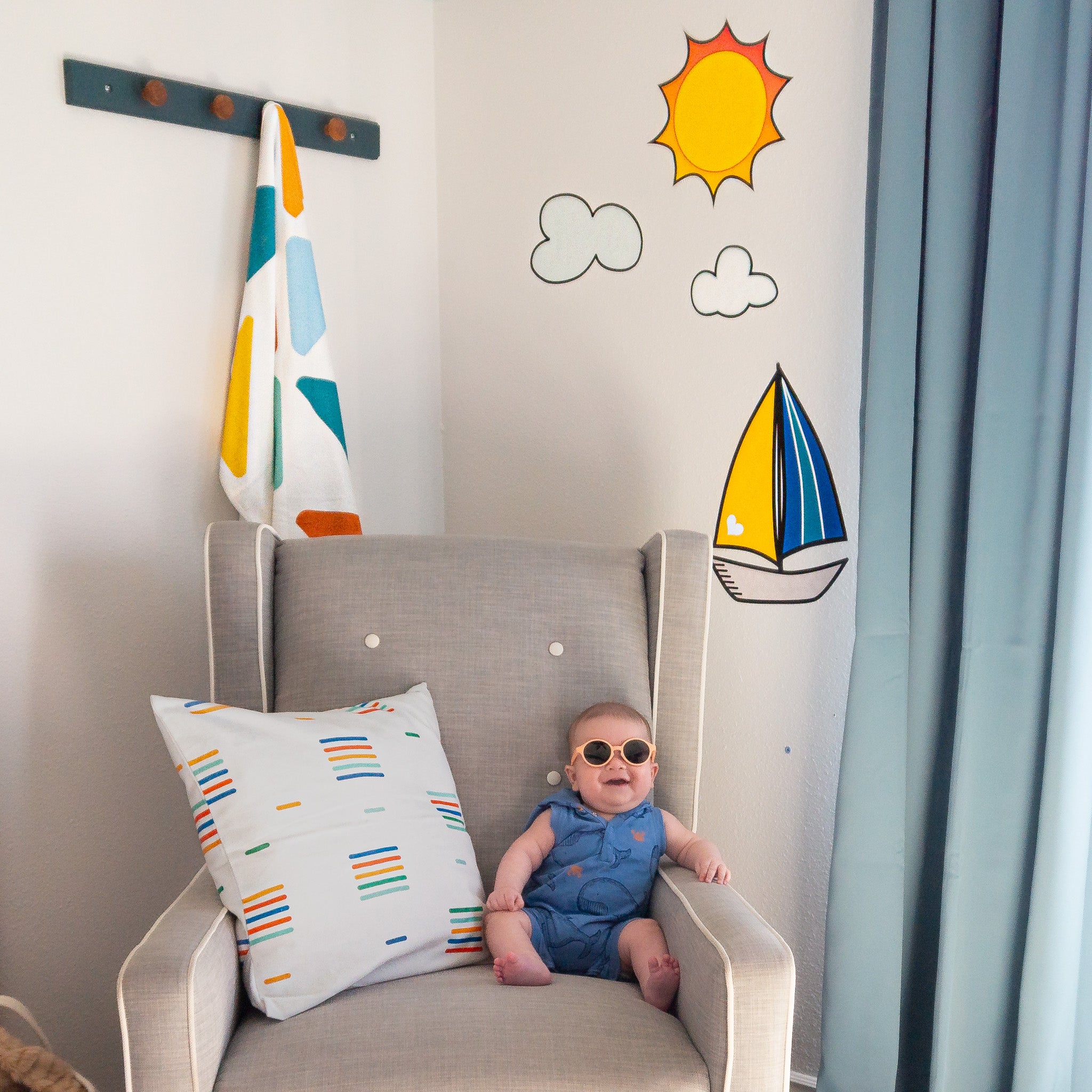 A baby sitting on a rocking chair wearing sunglasses with a colorful throw pillow next to him. On the wall behind there are wall decals of a sailboat, clouds and sun.