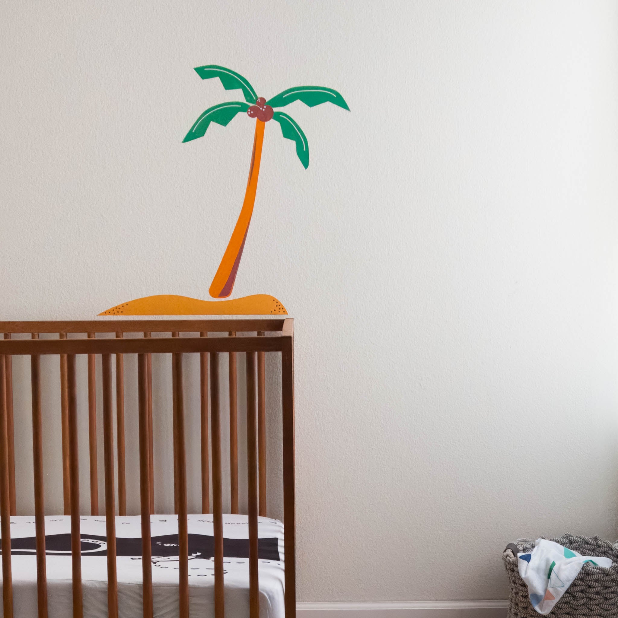 A white wall with a wall sticker of an island with a coconut tree. In front is a wood crib.