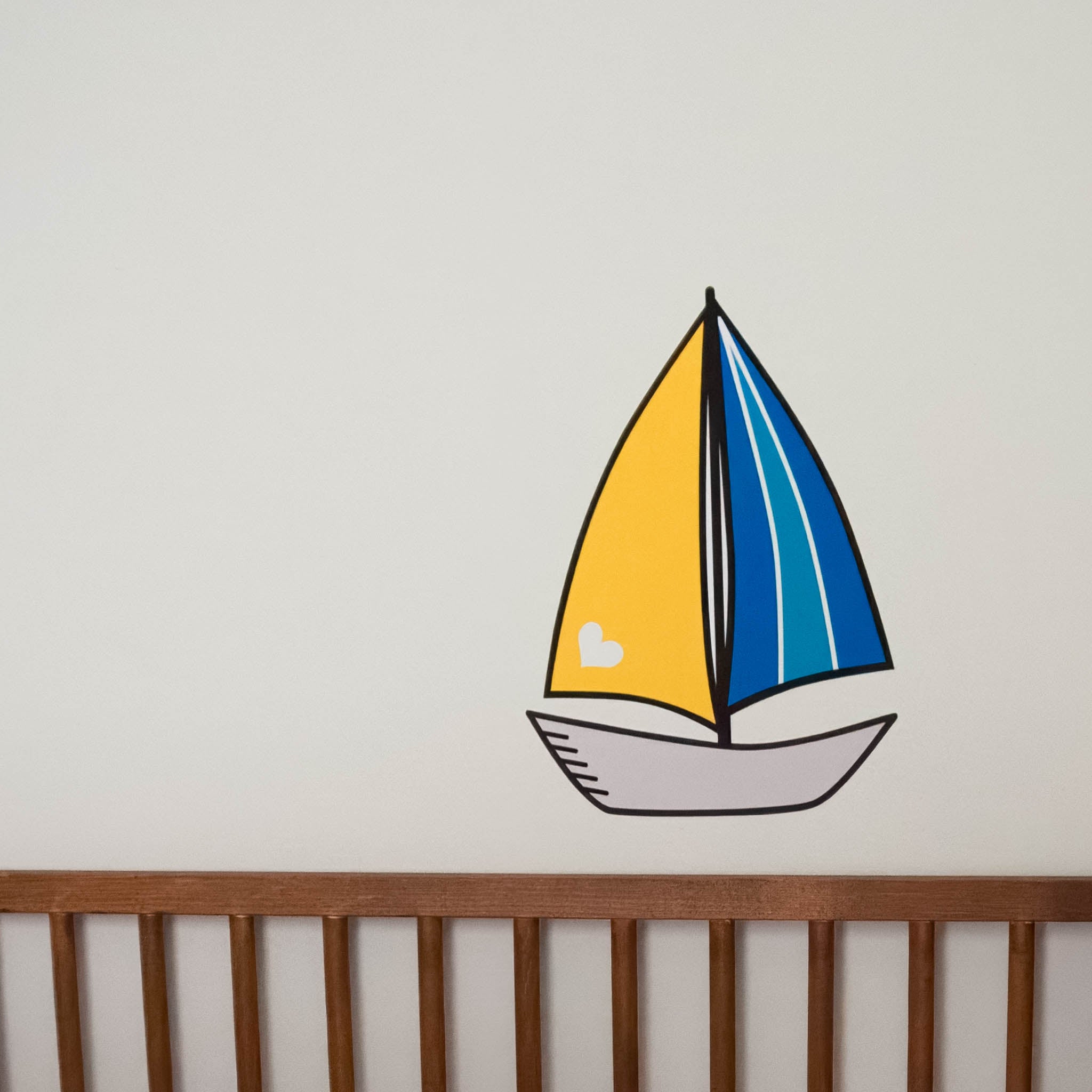 A crib rail in front of a white wall. On the white wall is a wall sticker of a sailboat with a yellow sail on the left and blue striped sail on the right.