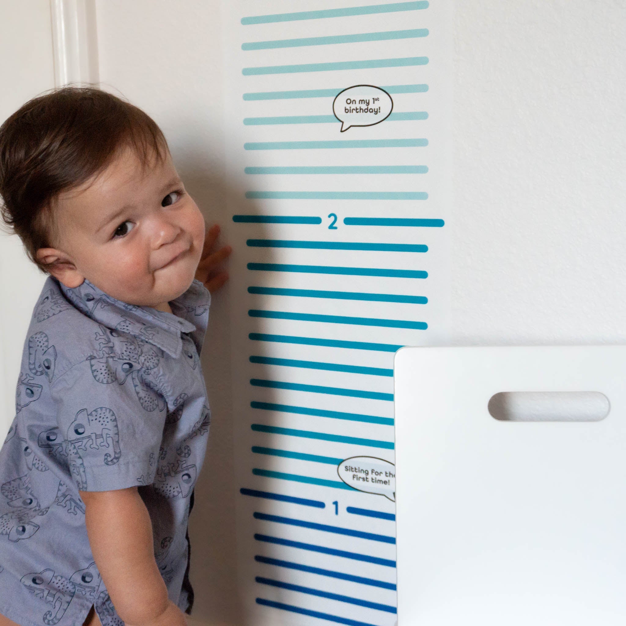 Toddler boy standing to the left of the Smartbaby Decor children's growth chart with add on sticker saying "1st birthday".