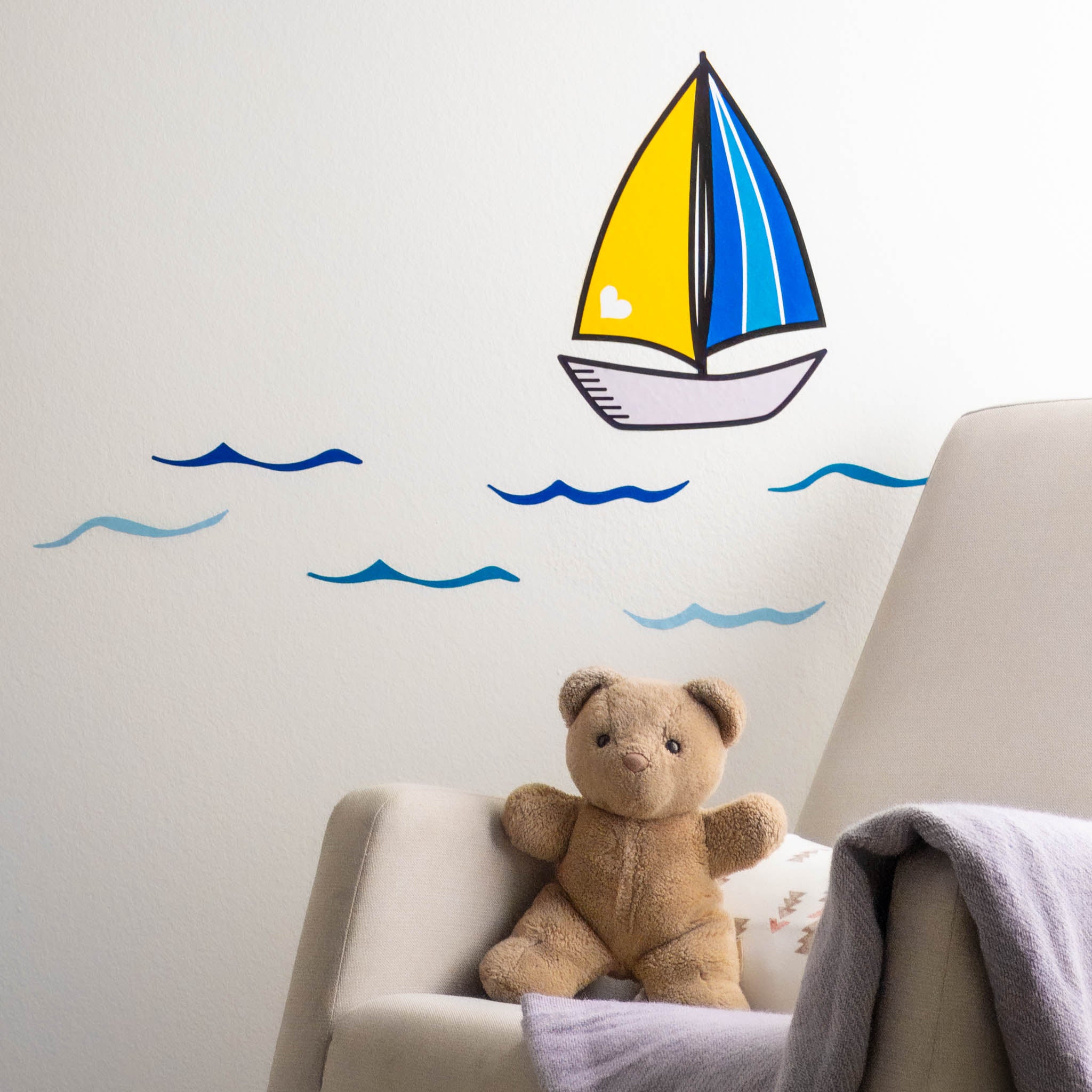 A wall with blue waves and sailboat wall stickers. A rocking chair with a teddy bear are to the right.