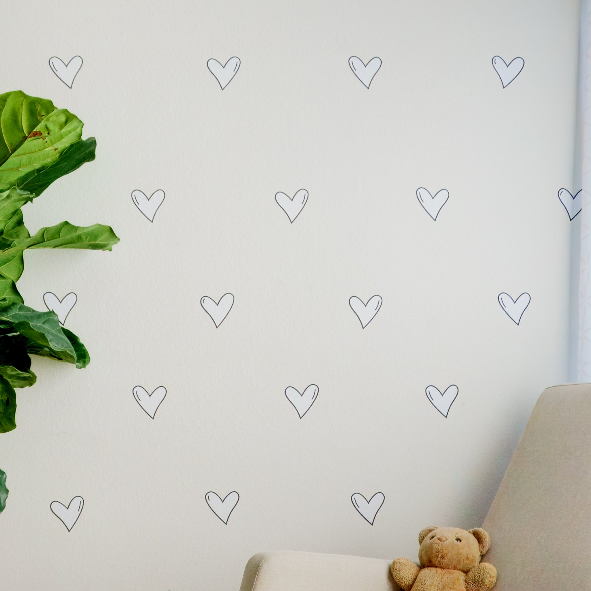 White wall with black and white heart wall decals arranged in a staggered pattern with a green plant on the left side and rocking chair with teddy bear on the right.