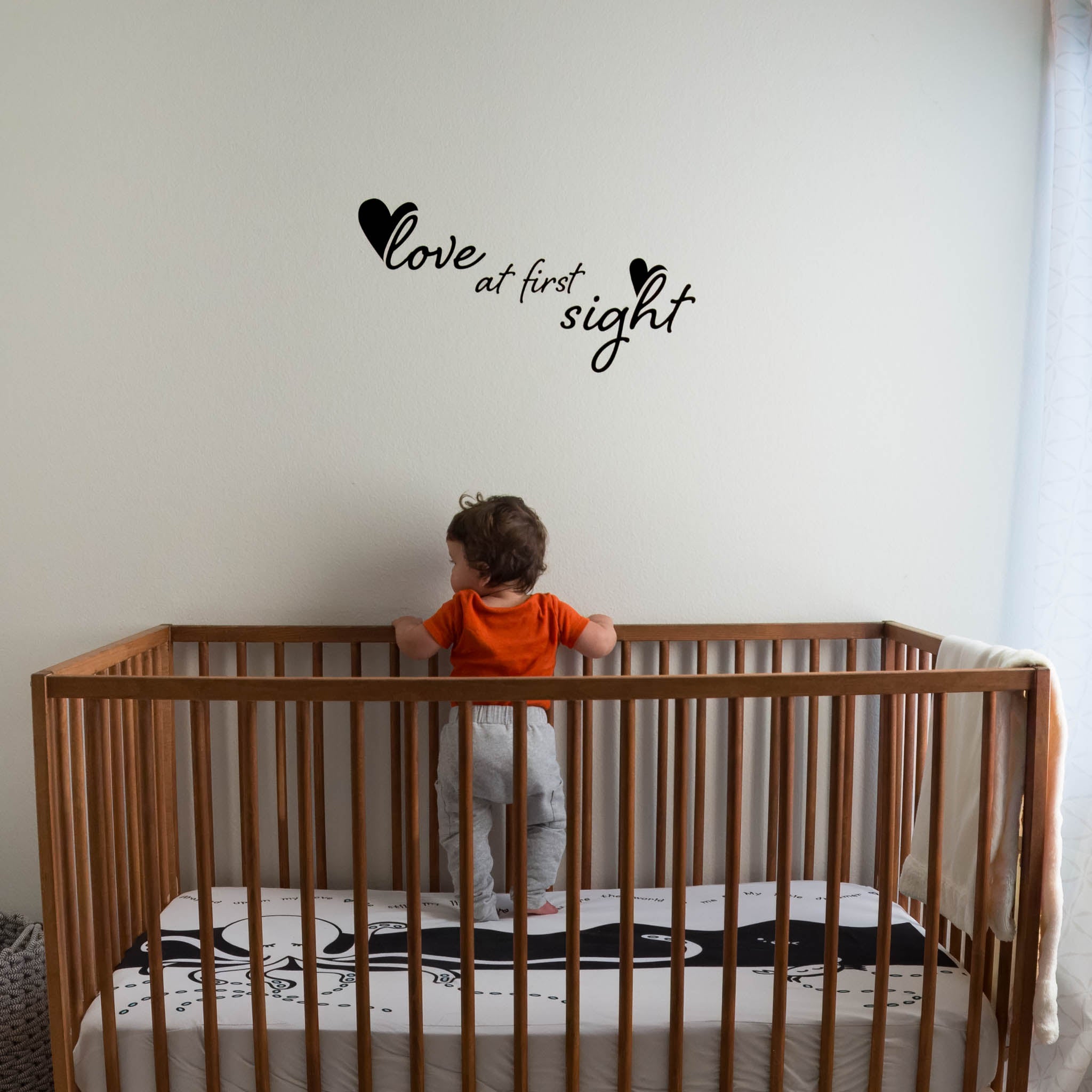 A baby standing in a wooden crib looking to the left side. The wall is white with the phrase "love at first sight" in black over his head.