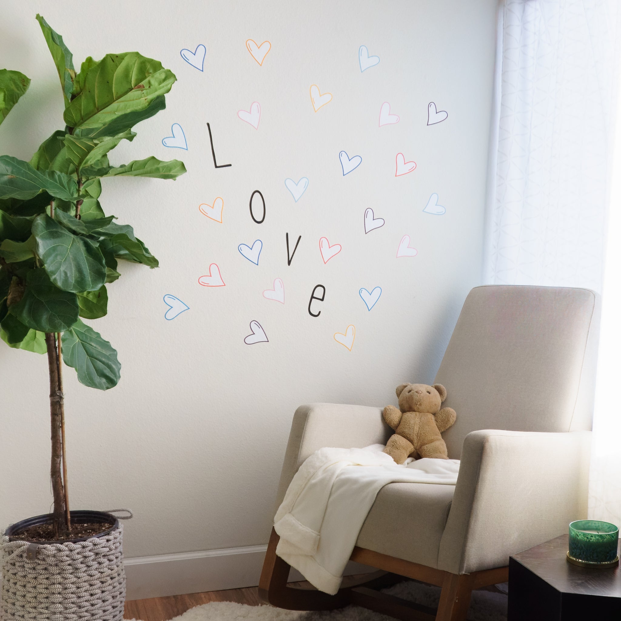 Nursery with green plant on left, rocking chair with teddy bear and blanket in front of a window on the right. On the wall by the rocking chair is the word "Love" in a black, modern font, surrounded by different colored heart wall decals. 