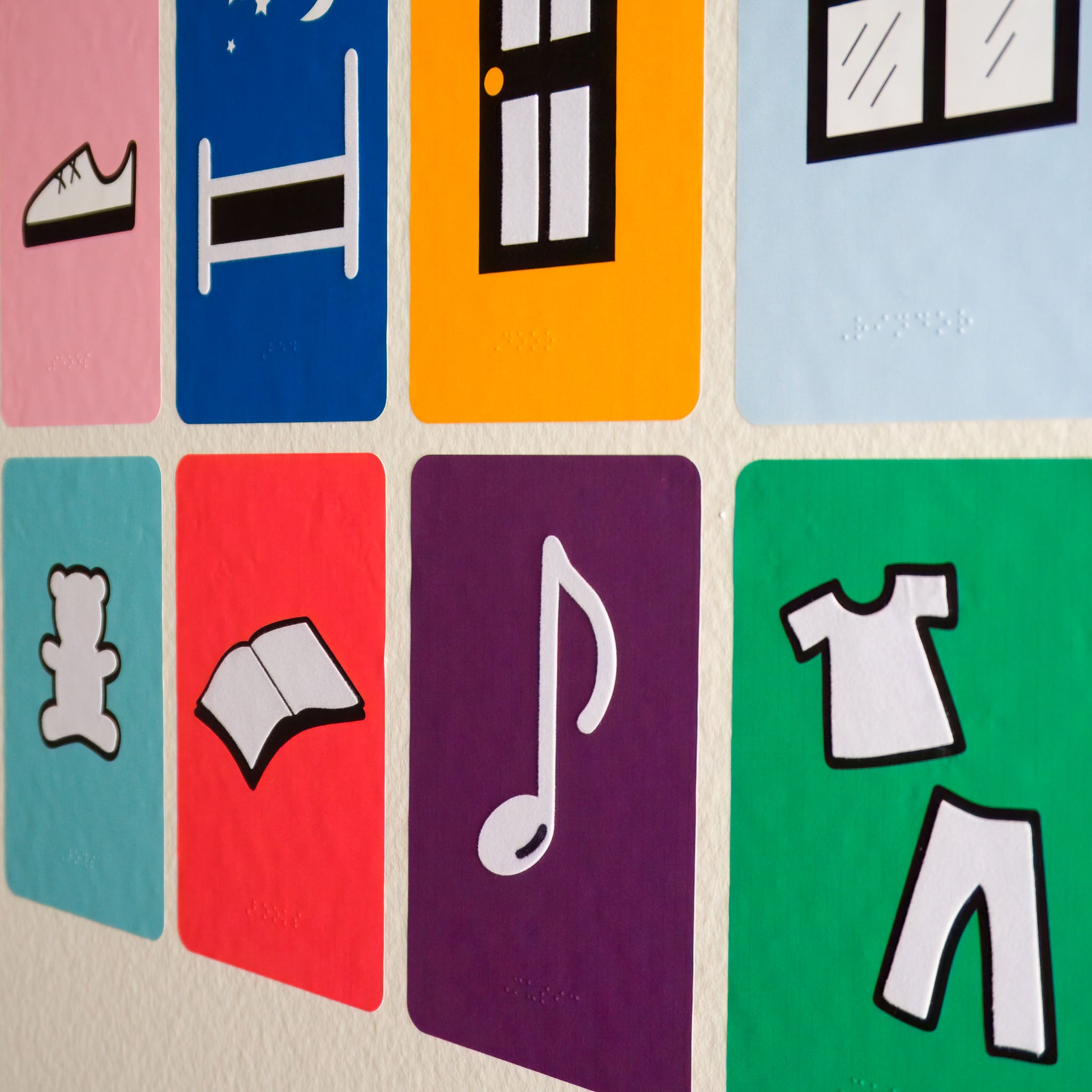 A photo of eight room labels for clothes, bed, door, window, toys, books, music and clothes with tactile components and braille