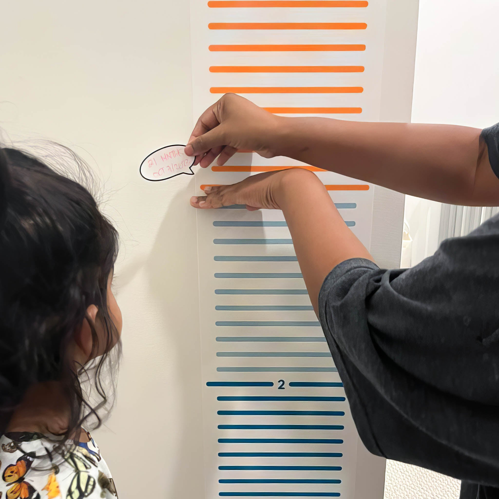 A colorful growth chart is on a white wall. A toddler girl is looking at it as her mother marks her height with a white sticker that reads "21 months".