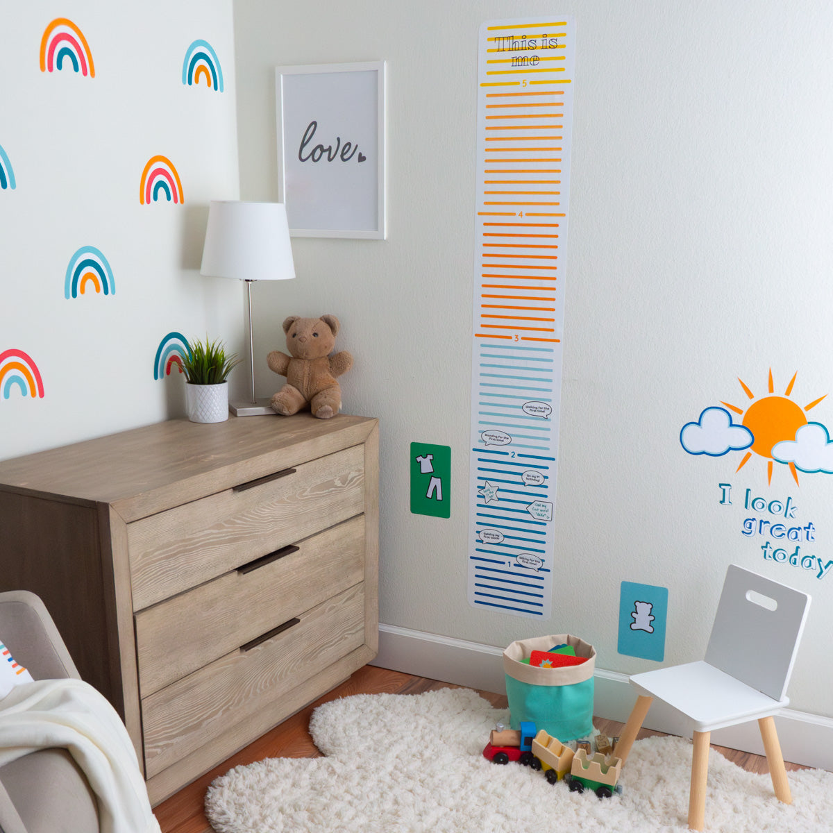 Child's nursery with a wall of scattered rainbow decals, colorful growth chart, teddy bear and lamp on a dresser with a child's chair and storage basket of toys