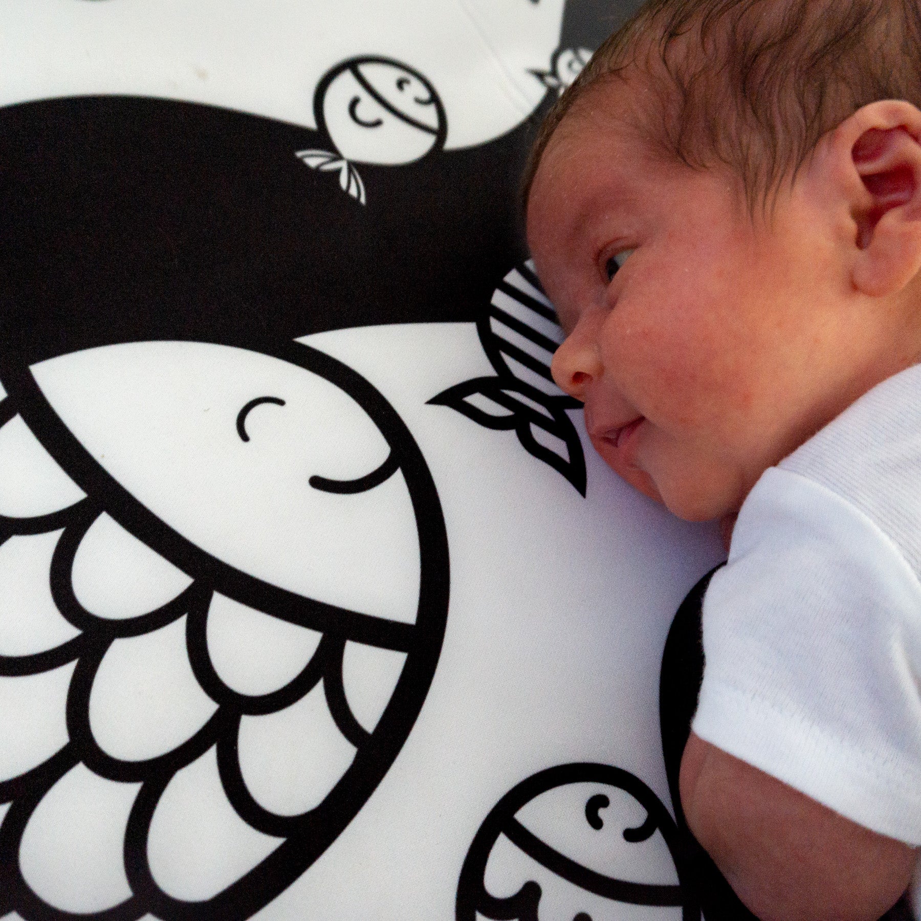 A newborn baby on the right laying on a mat with a print of fishes in black and whtie.