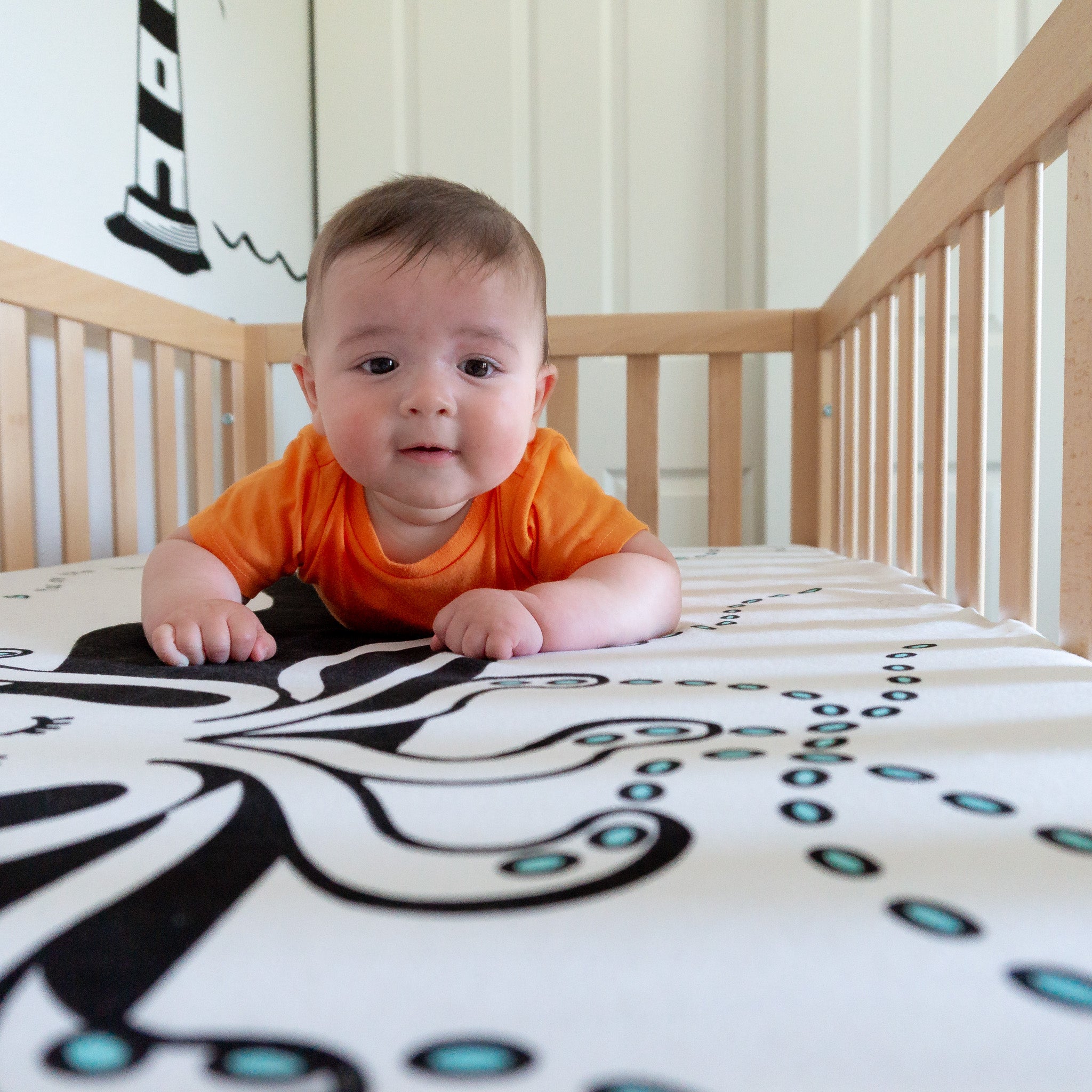 A baby wearing an orange shirt lying on a white and black crib sheet and smiling.