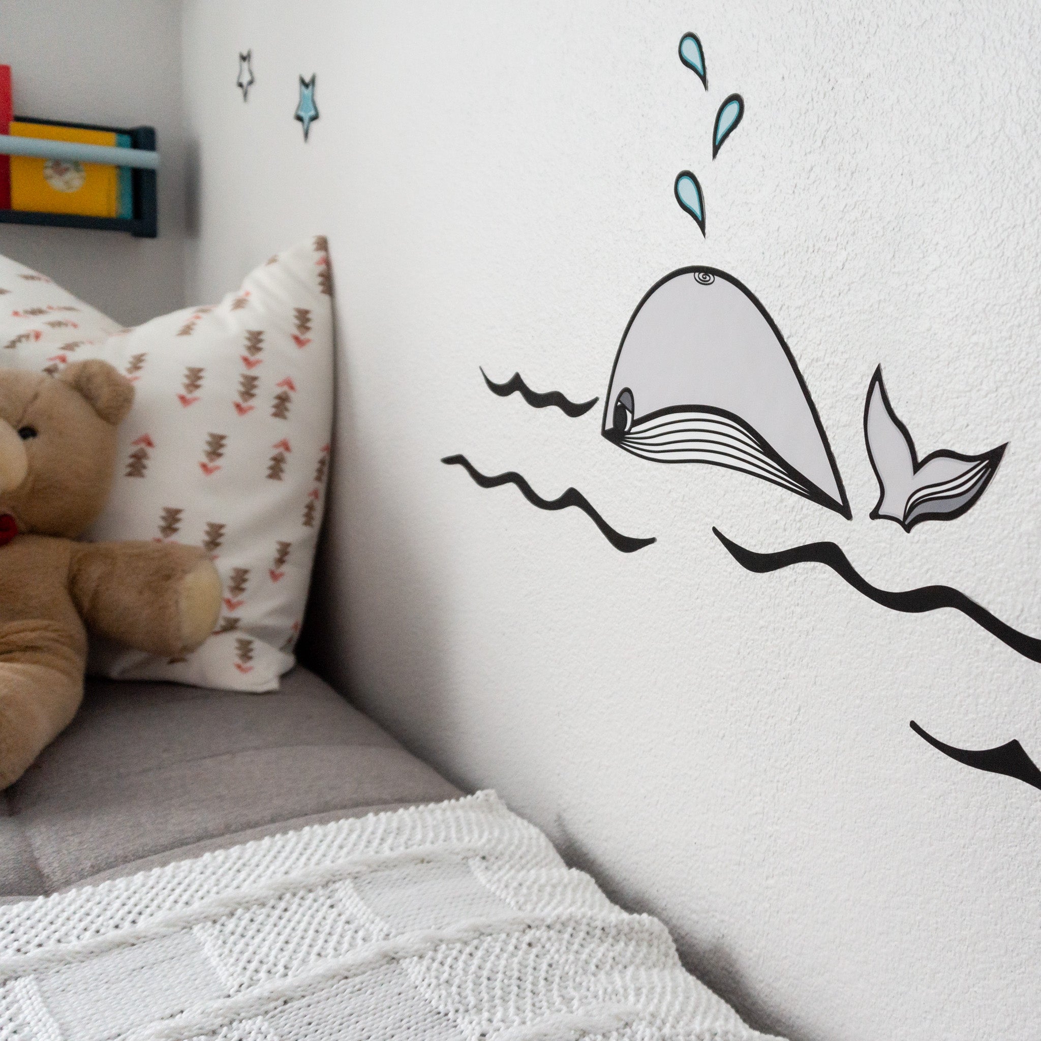 A corner of a child's room with a whale wall decal on the right side and a teddy bear and pillow on the right on a couch.