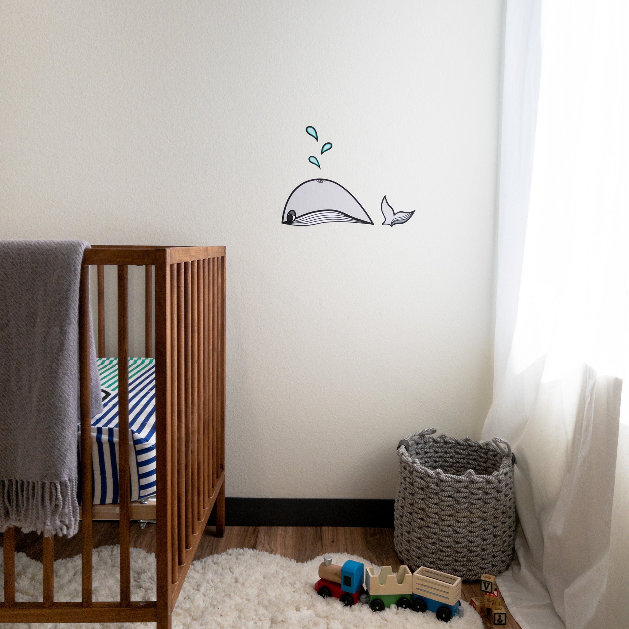 A baby's nursery with a white wall. On the white wall is a wall decal of a grey whale. On the floor is a rug with a toy train and there is a crib to the left.